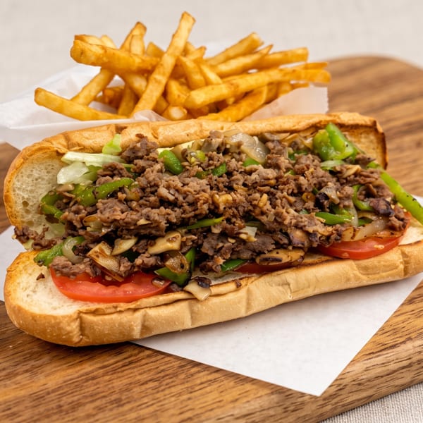 A cheesesteak sandwich with green peppers, onions, and mushrooms, served with fries on a wooden board.