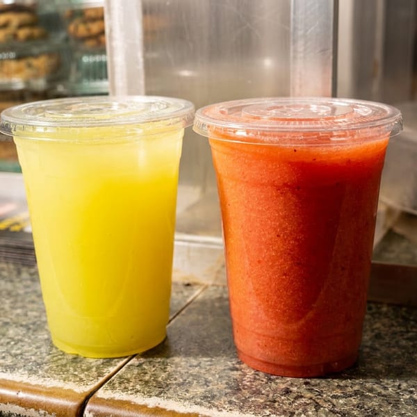 Yellow juice and red smoothie in plastic cups on a counter, with cookies in the background.