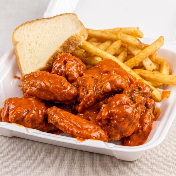 Spicy buffalo wings, french fries, and a slice of bread in a white takeout container.