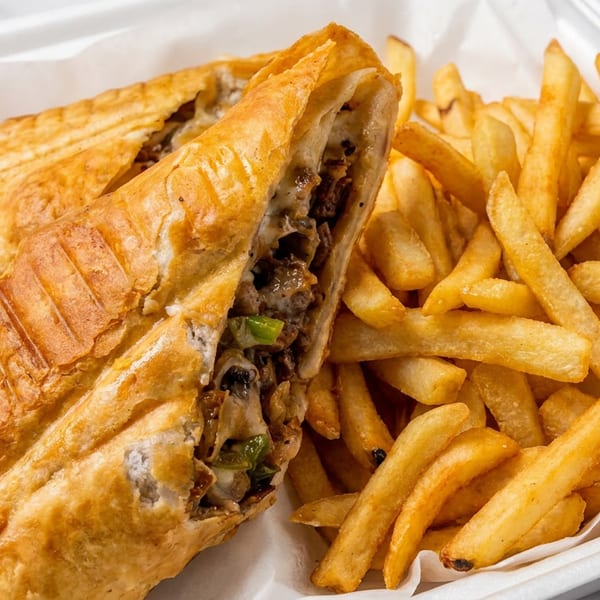 Two golden-brown cheesesteak pastries, one cut open, served with crispy french fries in a white container.